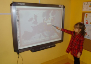 Dziewczynka stoi pod tablicą interaktywną i wskazuje na mapie Polskę.